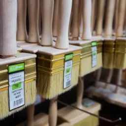 Rząd pędzli tapetowych z drewnianymi uchwytami i złotymi okuciami, ułożonych na półce w sklepie z artykułami budowlanymi, widoczne etykiety z numerami katalogowymi.