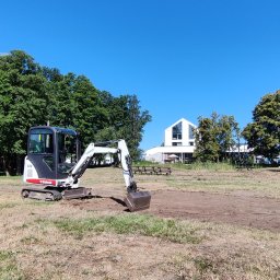 Dam-Kop - Mini koparka Bobcat na placu budowy przygotowanym pod wykop, w tle budynek z nowoczesną architekturą i zielone drzewa pod błękitnym niebem.