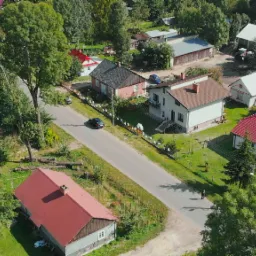 Zdjęcie z drona przedstawia wiejską scenerię z domami o r&oacute;żnej architekturze i kolorach dach&oacute;w, otoczonych zielenią drzew i trawnik&oacute;w, z widoczną drogą i przejeżdżającym samochodem.