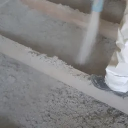 Aplikacja izolacji celulozowej na poddaszu za pomocą węża pneumatycznego, widoczne drewniane belki stropowe i pracownik w kombinezonie ochronnym.