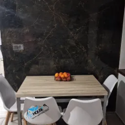 Jadalnia z drewnianym stołem, czterema białymi krzesłami z drewnianymi nogami i dekoracyjną ścianą w ciemnym marmurkowym wykończeniu. Na jednym z krzeseł widoczne logo firmy remontowej. Na stole...