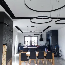 Nowoczesne wnętrze z sufitami podwieszanymi z okrągłymi lampami, widok na jadalnię z drewnianym stołem i czarnymi krzesłami, w tle kuchnia w odcieniach granatu, ściana z kominkiem i ułożonym drewnem.