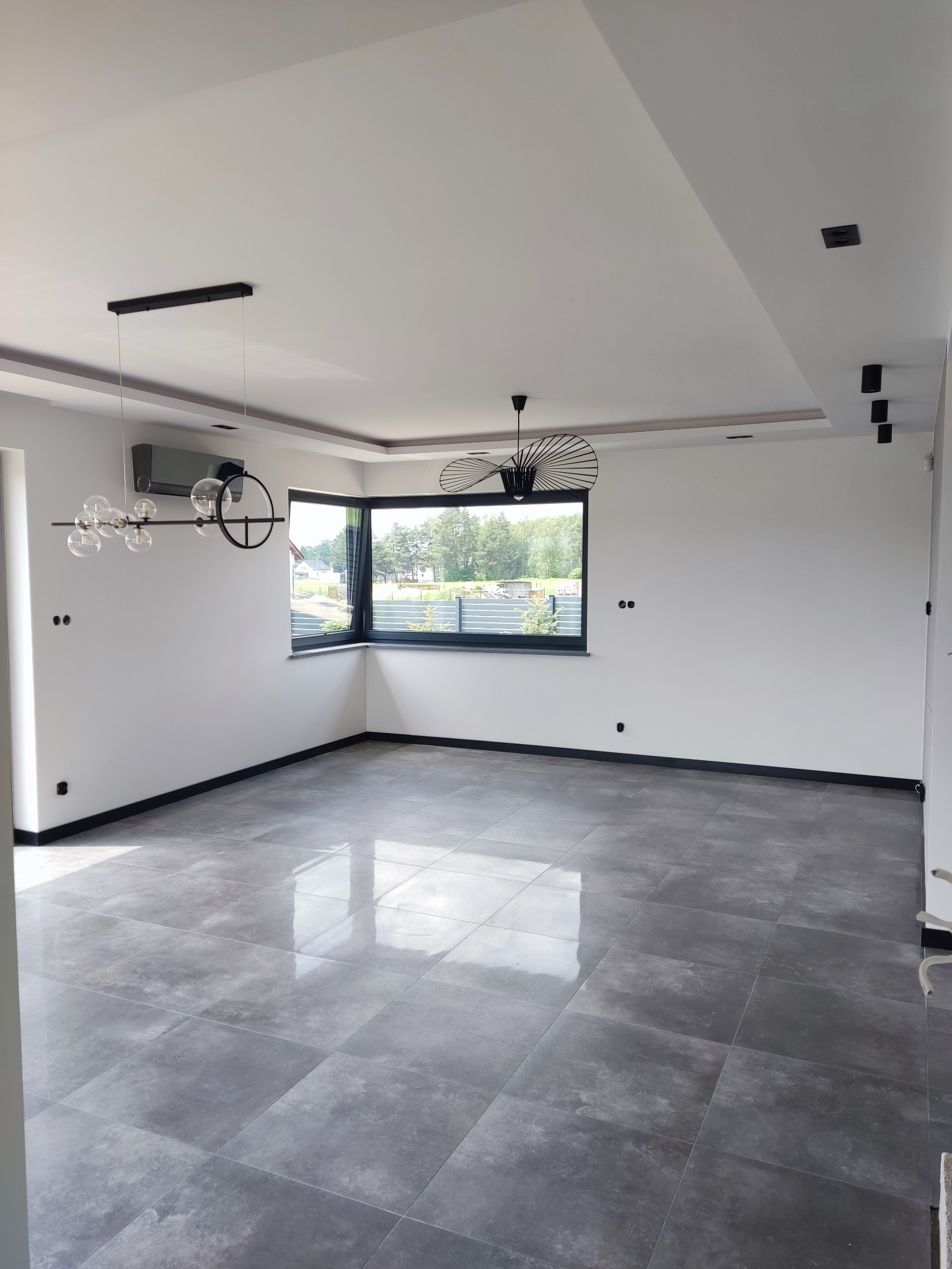 Nowoczesne wnętrze z szarymi płytkami podłogowymi, białymi ścianami, czarnymi listwami przypodłogowymi i designerskim oświetleniem: lampa wisząca z szklanymi kulami i lampa sufitowa z metalowymi...