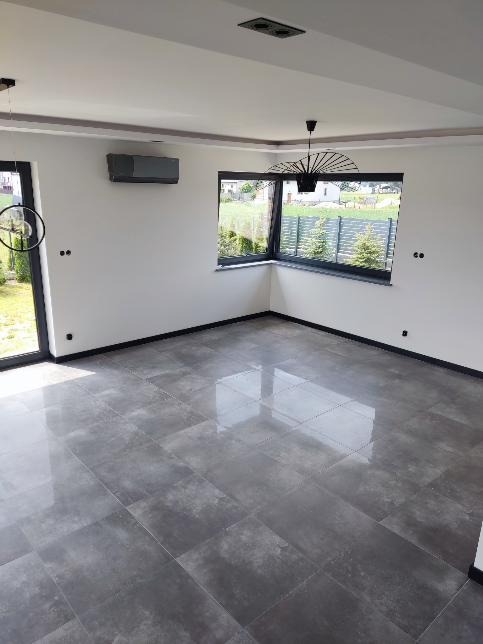Puste, jasne pomieszczenie z szarą, błyszczącą podłogą z płytek, dużymi oknami z widokiem na ogród i nowoczesnym oświetleniem sufitowym.