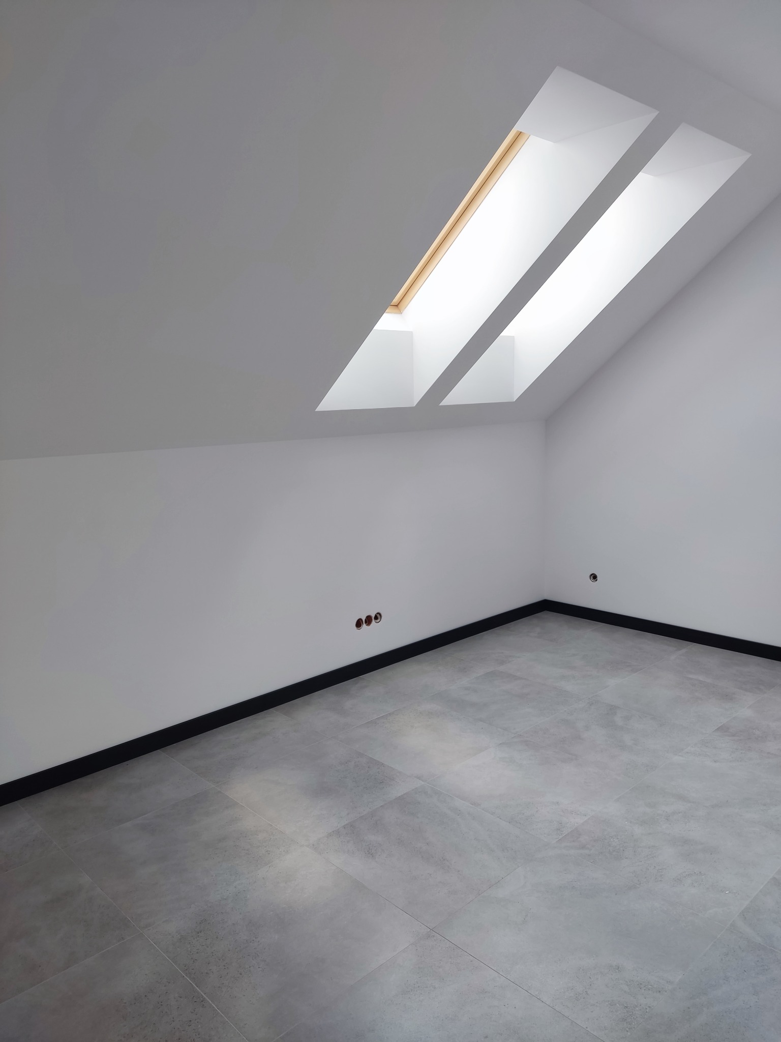 Poddasze po remoncie z dwoma oknami dachowymi, szarą podłogą z płytek i białymi ścianami z widocznymi gniazdkami elektrycznymi.