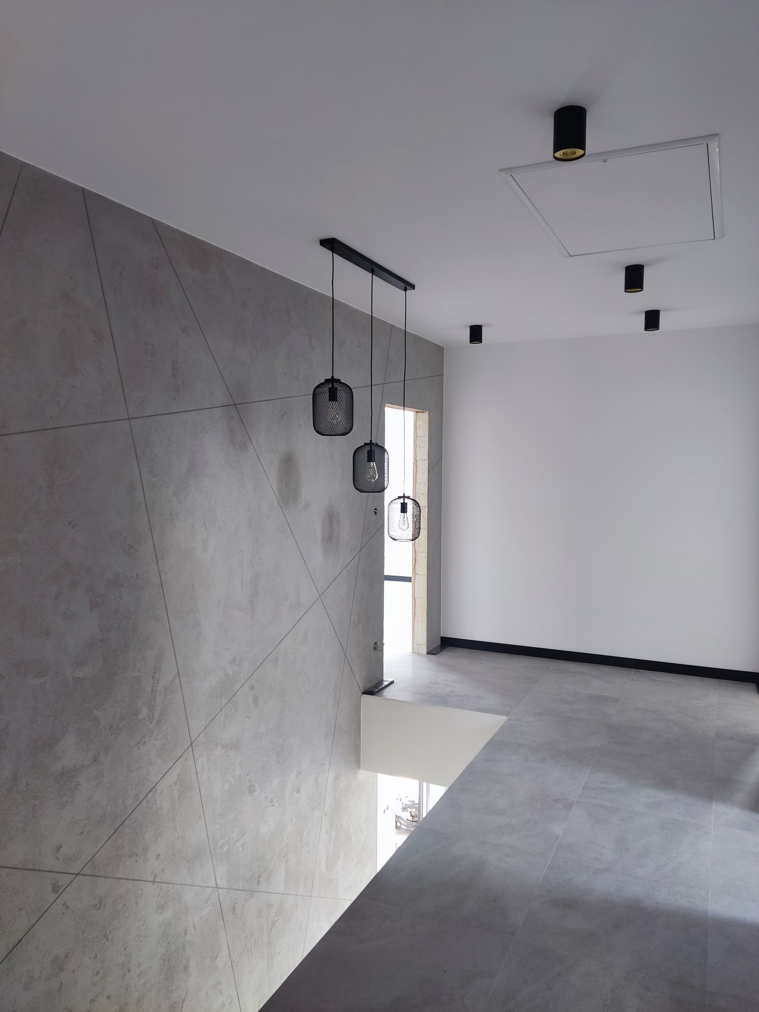 Nowoczesne wnętrze: szara ściana z geometrycznym wzorem, wiszące lampy z metalowymi kloszami, biała ściana z czarnymi punktowymi lampami, szara podłoga z płytek.