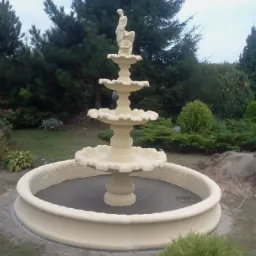 Trójpoziomowa fontanna ogrodowa z figurą na szczycie, otoczona zielenią i świeżo posianą trawą.