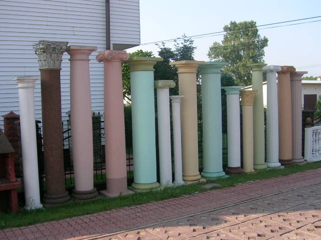 Różnokolorowe kolumny betonowe o różnych wzorach i wysokościach ustawione w rzędzie na trawie obok chodnika z kostki, na tle budynku z białym sidingiem i ogrodzeniem.