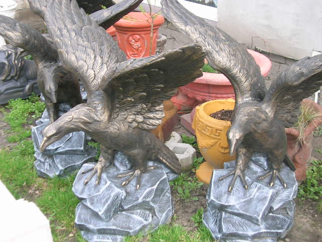 Brązowe figury orłów z rozpostartymi skrzydłami, ustawione na postumentach imitujących skały, otoczone donicami i zielenią w ogrodzie.