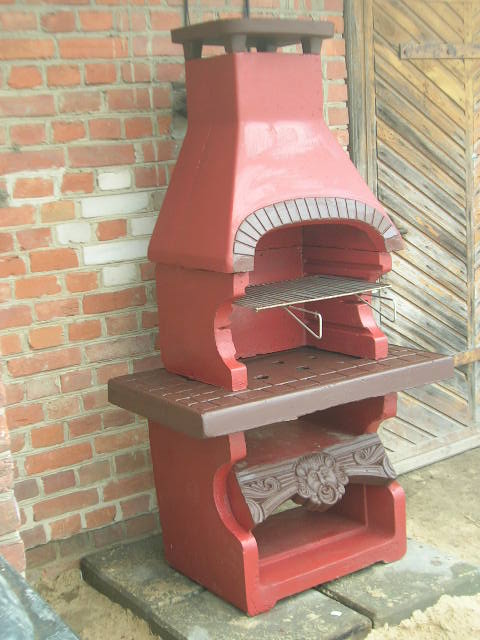 Czerwono-brązowy, betonowy grill ogrodowy z dekoracyjnym motywem lwa, ustawiony na betonowych płytach przed ceglaną ścianą i drewnianymi drzwiami.