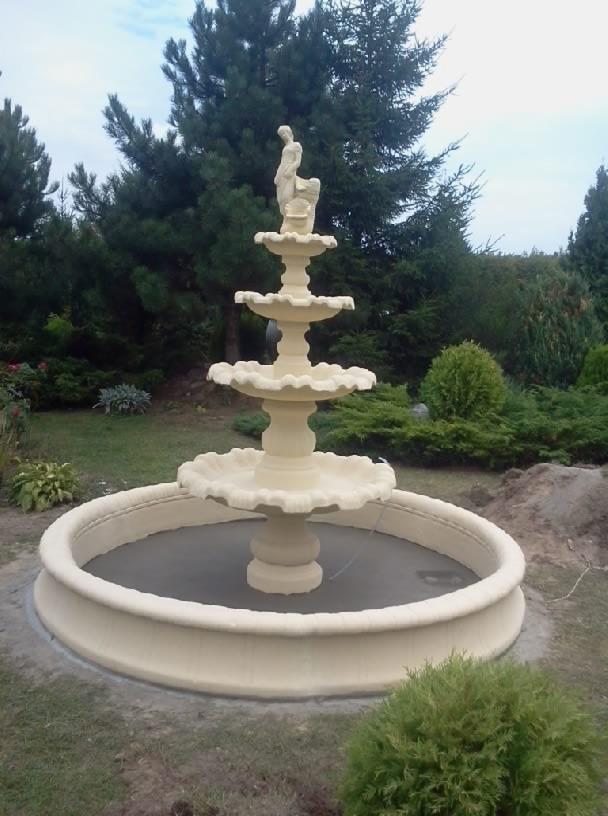 Trójpoziomowa fontanna ogrodowa z figurą na szczycie, otoczona zielenią i świeżo posianą trawą.