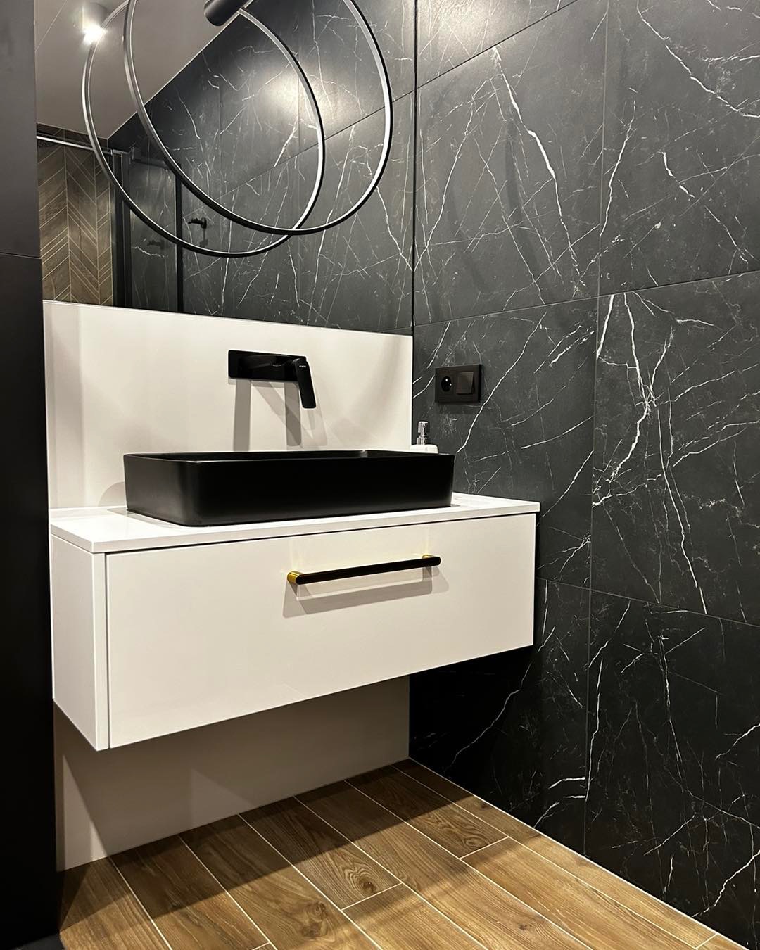 Nowoczesna łazienka z czarną umywalką nablatową i armaturą na tle płytek imitujących czarny marmur z białymi żyłami, drewnopodobna podłoga, okrągłe lustro z podświetleniem.