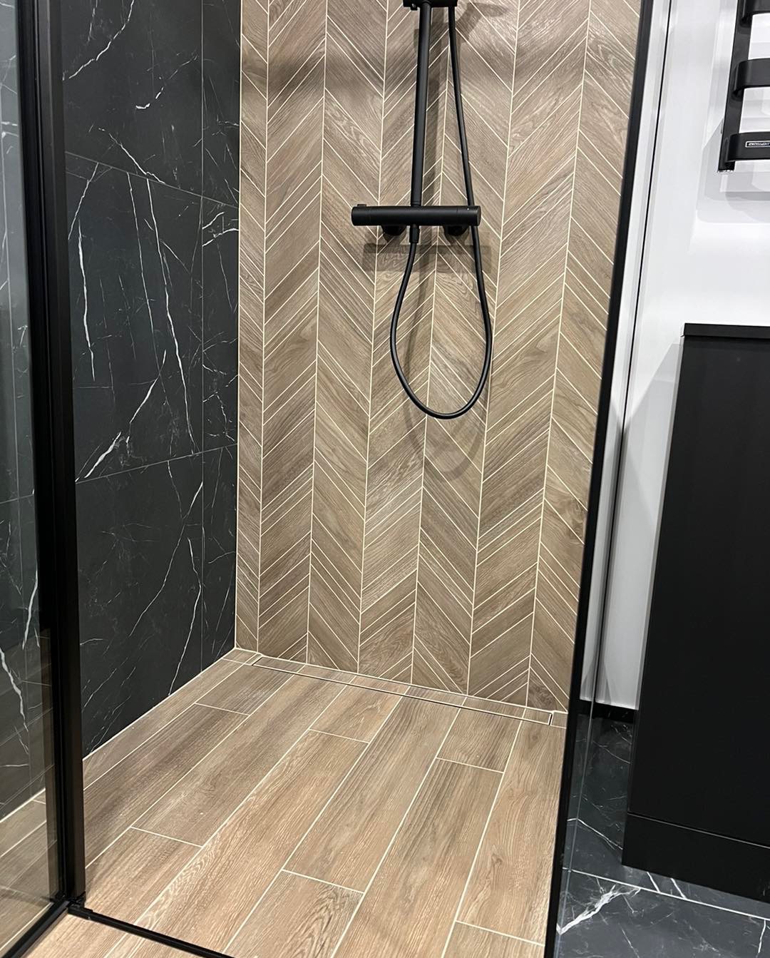 Nowoczesny prysznic walk-in z czarną armaturą i płytkami imitującymi drewno ułożonymi w jodełkę na ścianie, oraz czarnymi płytkami imitującymi marmur na bocznej ścianie.