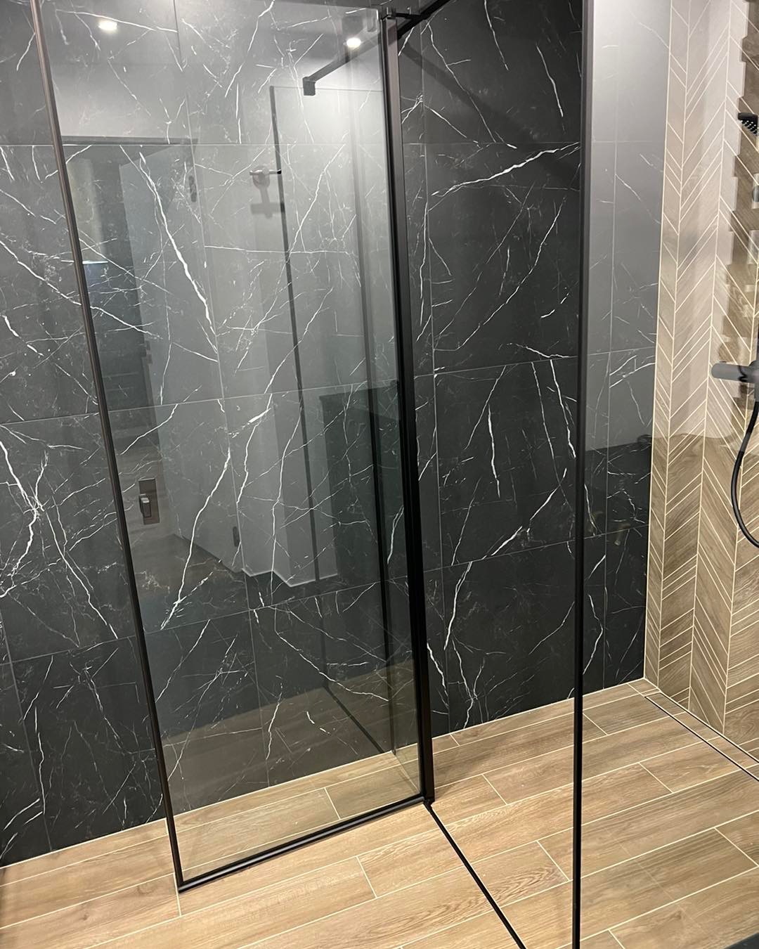 Nowoczesna kabina prysznicowa z czarnymi płytkami imitującymi marmur i drewnopodobnymi płytkami podłogowymi ułożonymi w jodełkę na ścianie.