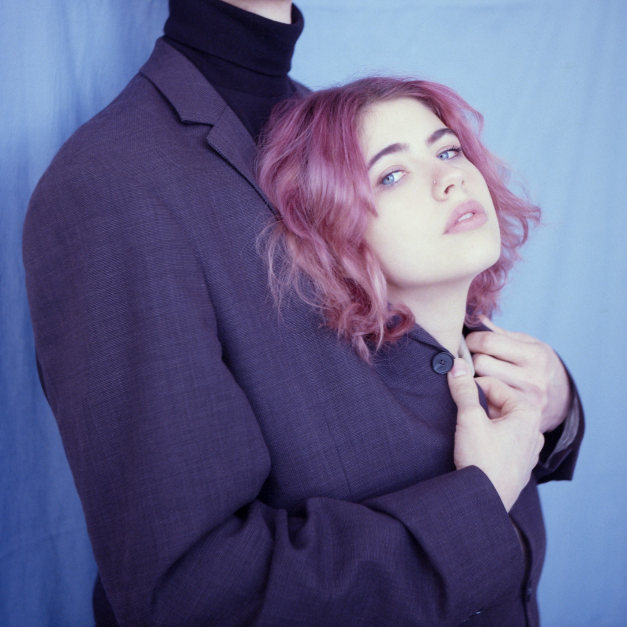 Portret kobiety z różowymi włosami, opierającej głowę na ramieniu mężczyzny w ciemnym garniturze na tle błękitnej tkaniny.