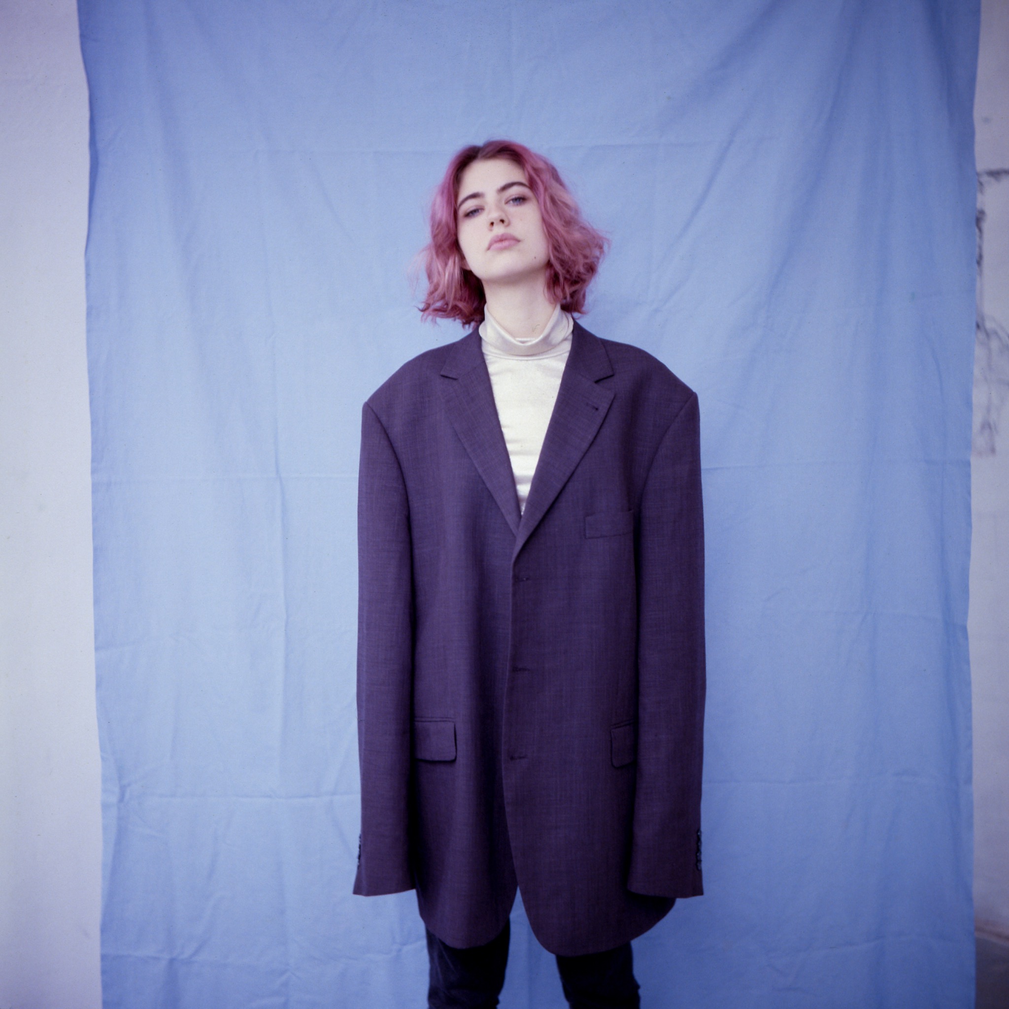 Portret młodej kobiety z różowymi włosami, ubrana w za dużą, fioletową marynarkę na tle błękitnego płótna.