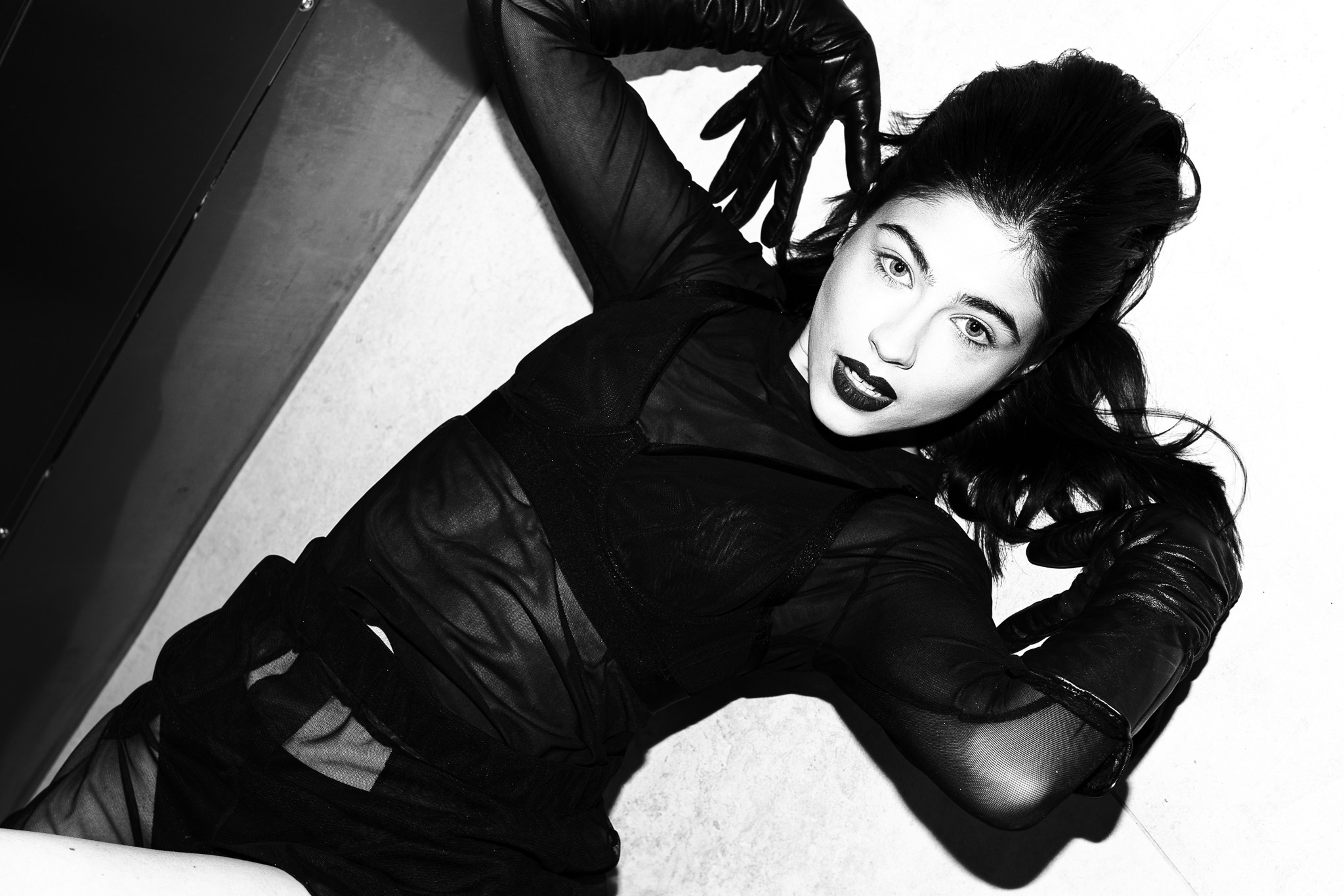 Czarno-białe zdjęcie kobiety w czarnej, przezroczystej bluzce, skórzanych rękawiczkach i z mocnym makijażem, leżącej na białym tle i patrzącej w obiektyw.