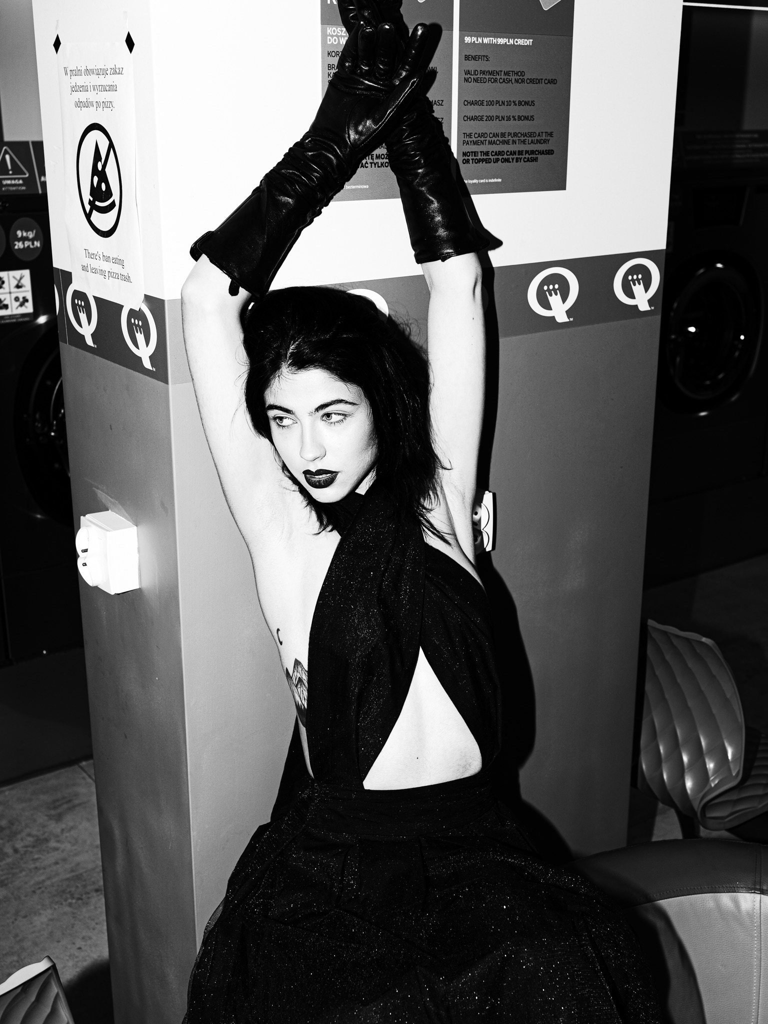 Kobieta w czarnej sukni i długich rękawicach pozuje na tle ściany z informacjami o pralni samoobsługowej, z widocznym znakiem zakazu spożywania pizzy.