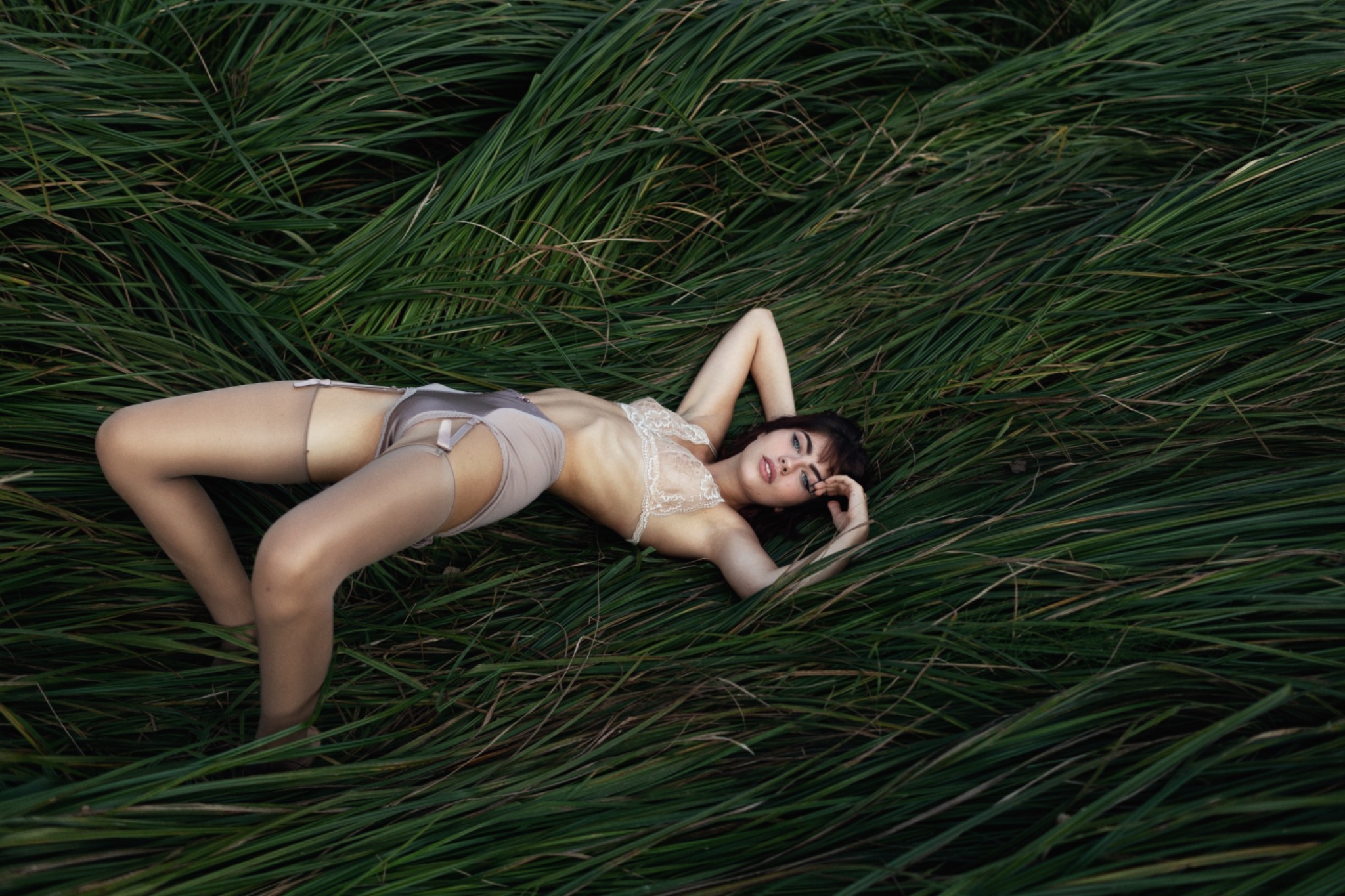 Kobieta w bieliźnie z koronką i pończochami leży na łące wysokiej trawy, spoglądając w obiektyw