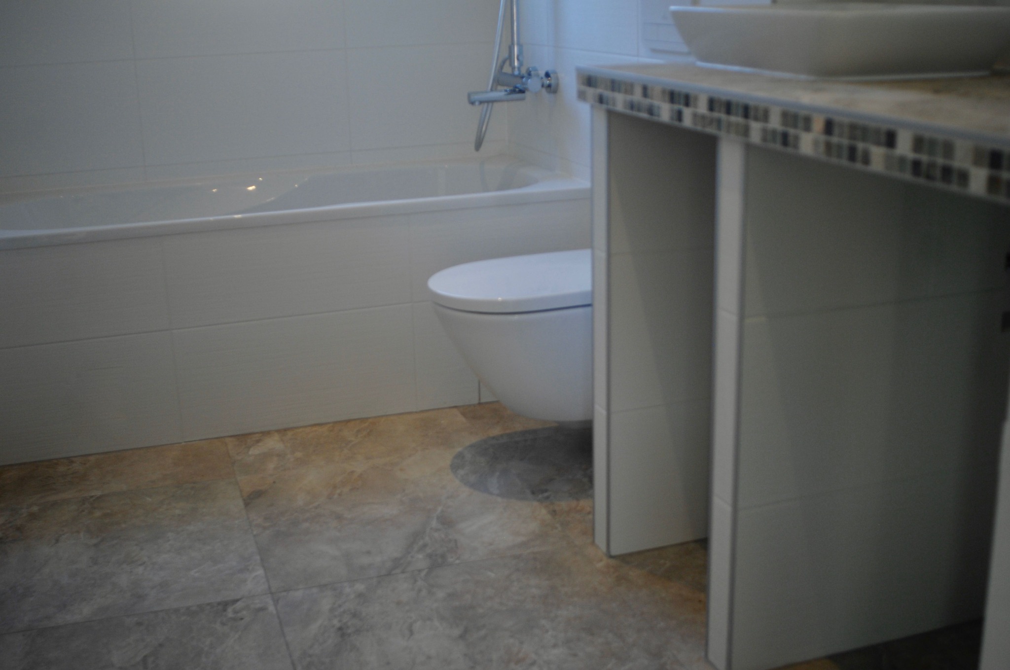 Nowoczesna łazienka z białą armaturą, wanną i toaletą typu Geberit, wykończona płytkami imitującymi marmur na podłodze, z blatem umywalkowym wykończonym mozaiką.