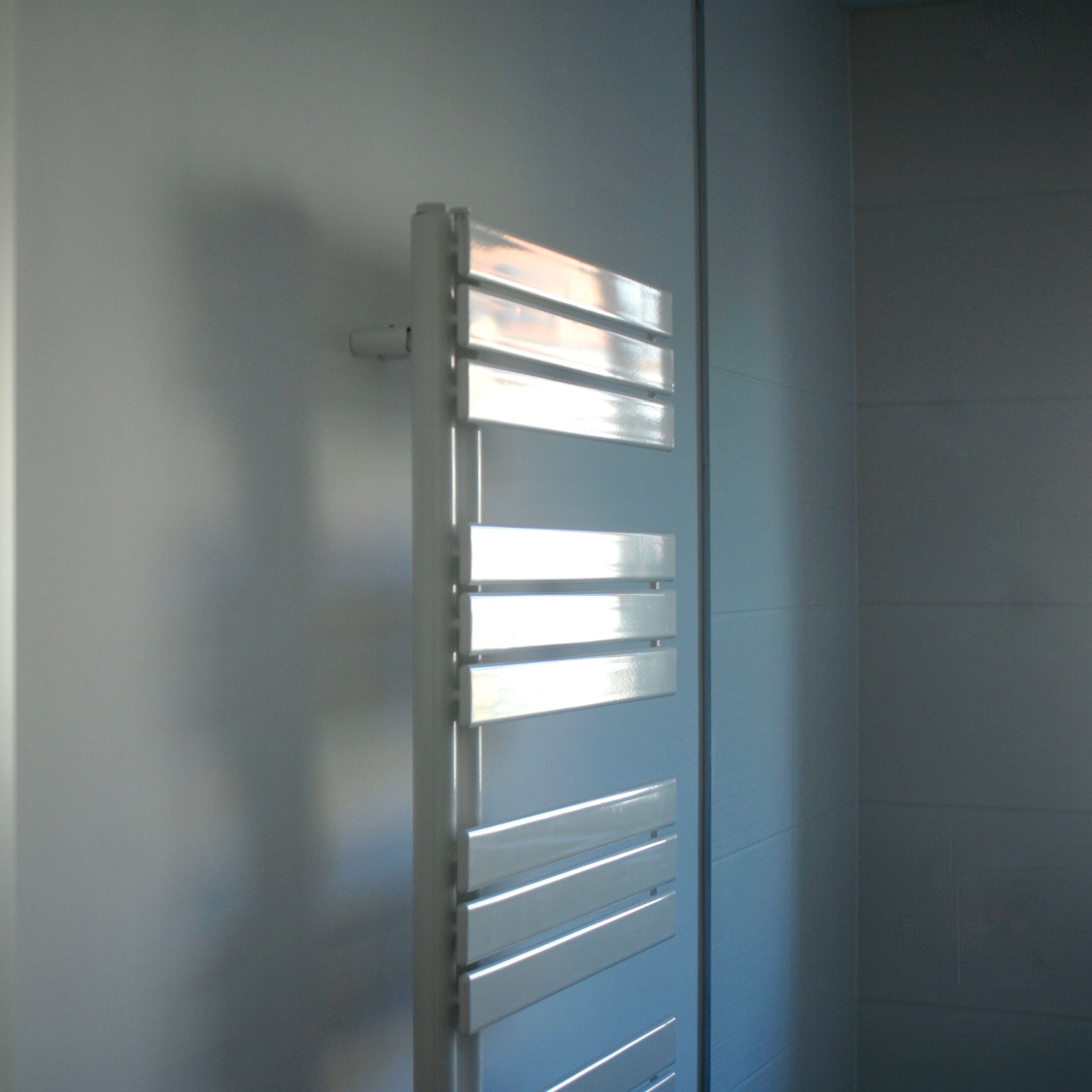 Biały grzejnik drabinkowy zamontowany na jasnej, kafelkowanej ścianie w łazience, oświetlony bocznym światłem słonecznym.