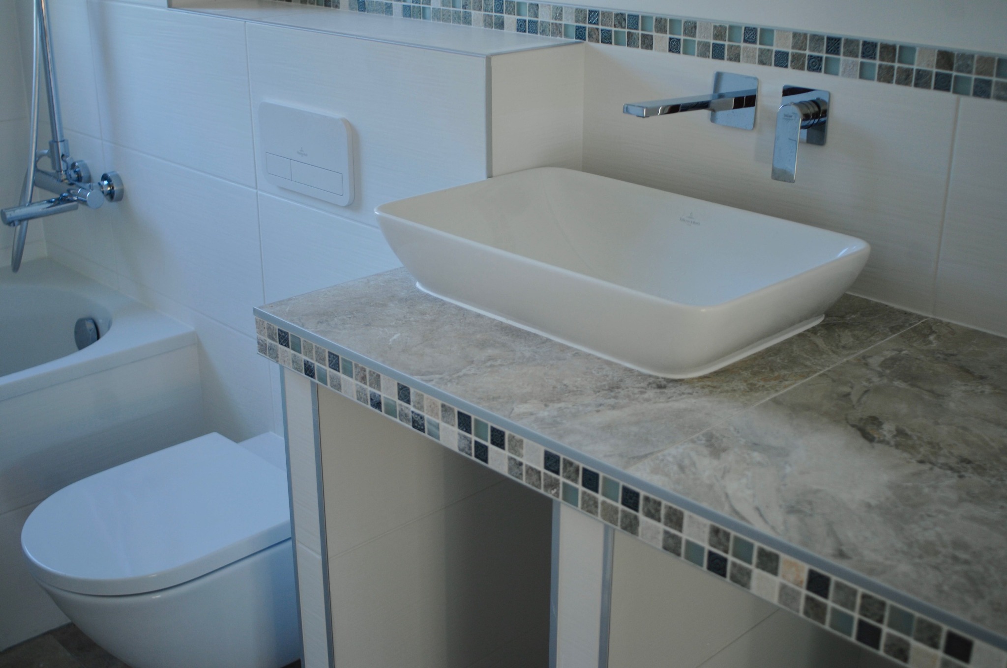 Nowoczesna łazienka z umywalką nablatową o prostokątnym kształcie, baterią ścienną, podwieszaną toaletą i wanną, wykończona białymi płytkami i mozaiką.