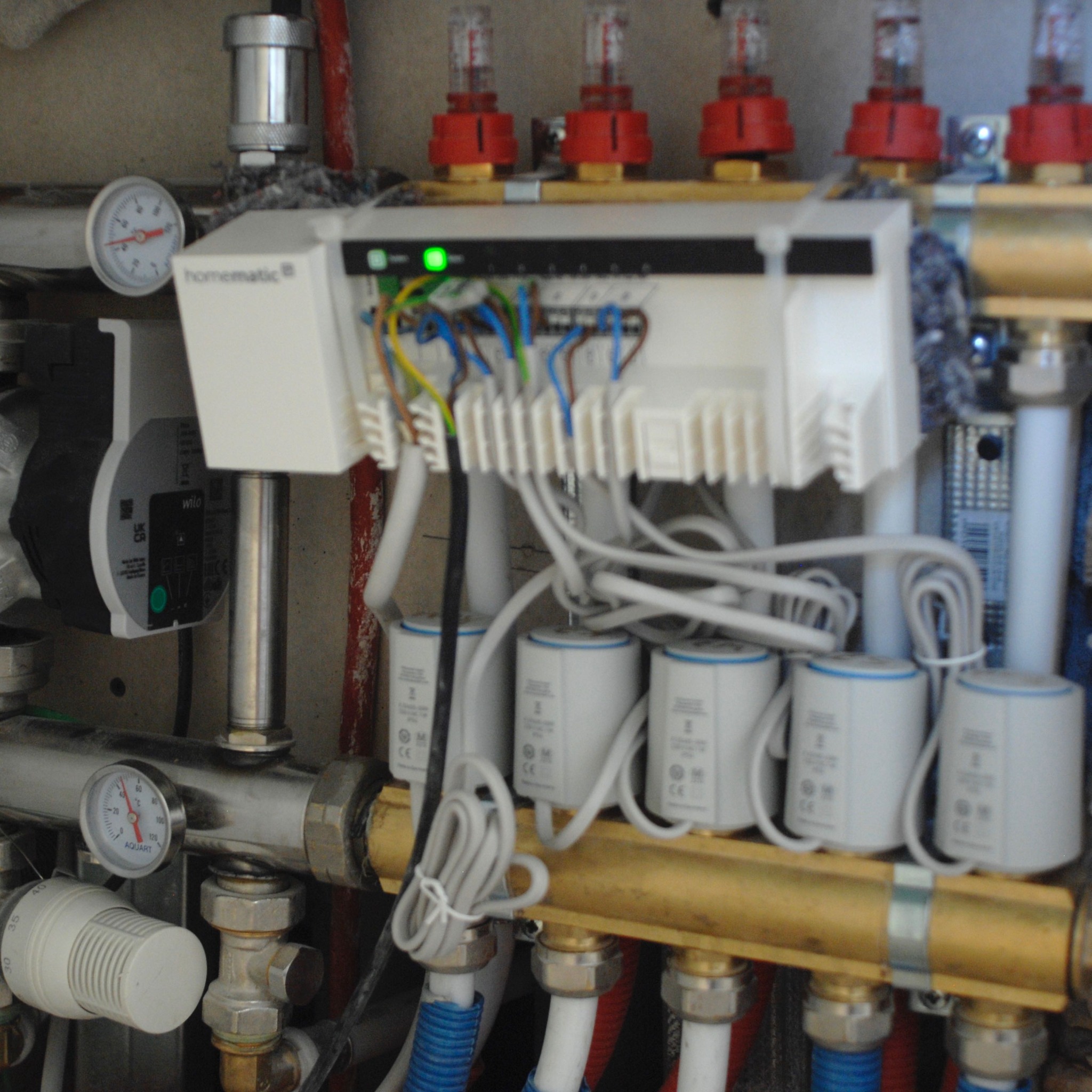 Rozdzielacz ogrzewania podłogowego z widocznymi zaworami termoelektrycznymi, przewodami, manometrami i modułem sterującym Homematic z zieloną diodą LED.