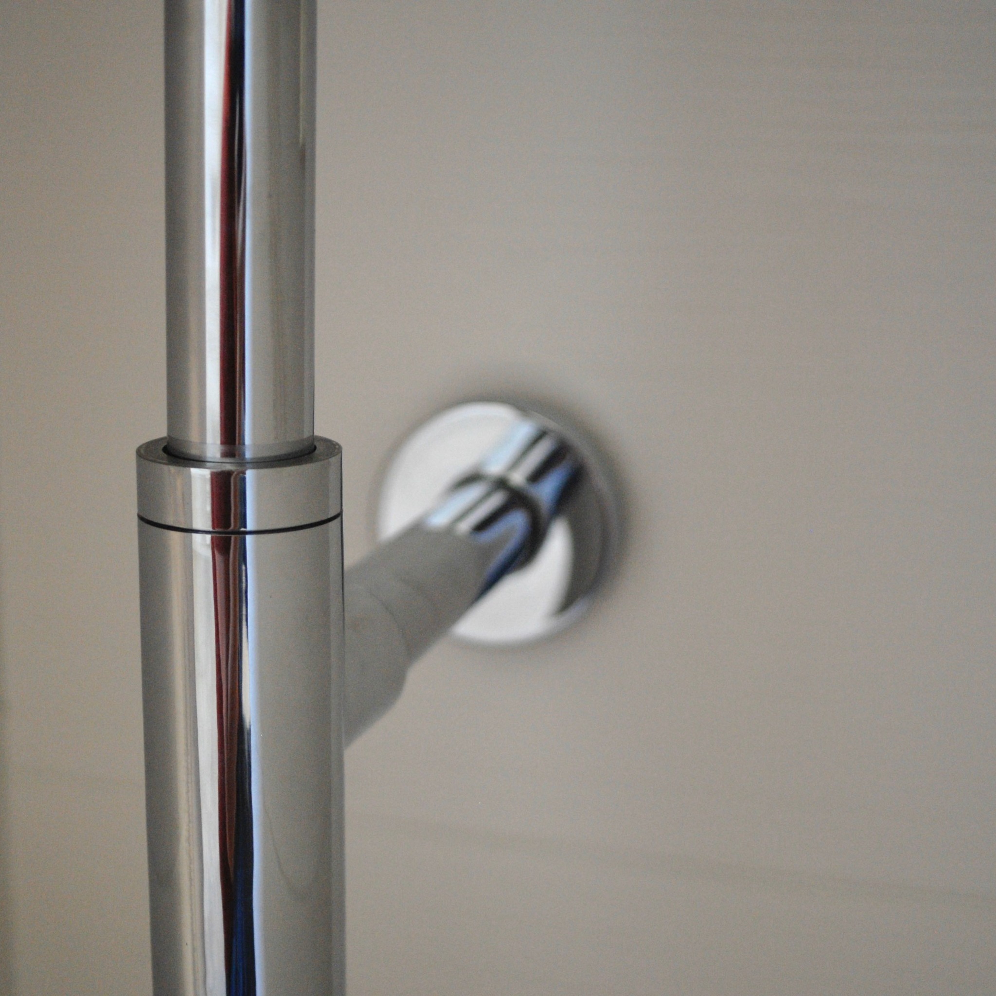 Błyszcząca chromowana rura pionowa z fragmentem poziomej rury przy ścianie w łazience, widoczne odbicia światła.