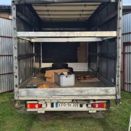 Wnętrze paki busa z otwartą plandeką, widoczne kartony, opony i metalowa skrzynka, na podłodze leżą pasy mocujące, widok z tyłu, tablica rejestracyjna KPR 5K30.