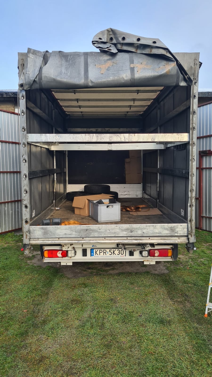 Wnętrze paki busa z otwartą plandeką, widoczne kartony, opony i metalowa skrzynka, na podłodze leżą pasy mocujące, widok z tyłu, tablica rejestracyjna KPR 5K30.