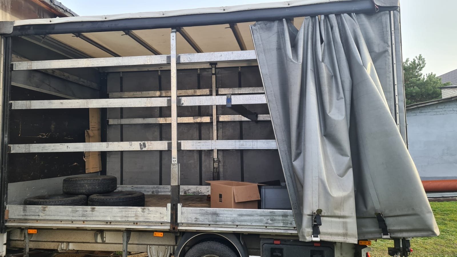 Wnętrze skrzyni ładunkowej busa z częściowo odsłoniętą plandeką, widoczne trzy opony i kartonowe pudło w środku, szare ściany i metalowe wzmocnienia.