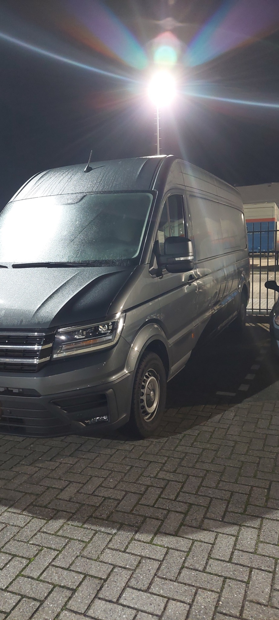 Szary bus marki Volkswagen Crafter zaparkowany na brukowanym parkingu nocą, oświetlony przez jasną lampę uliczną, widoczne krople wody na masce i szybie.