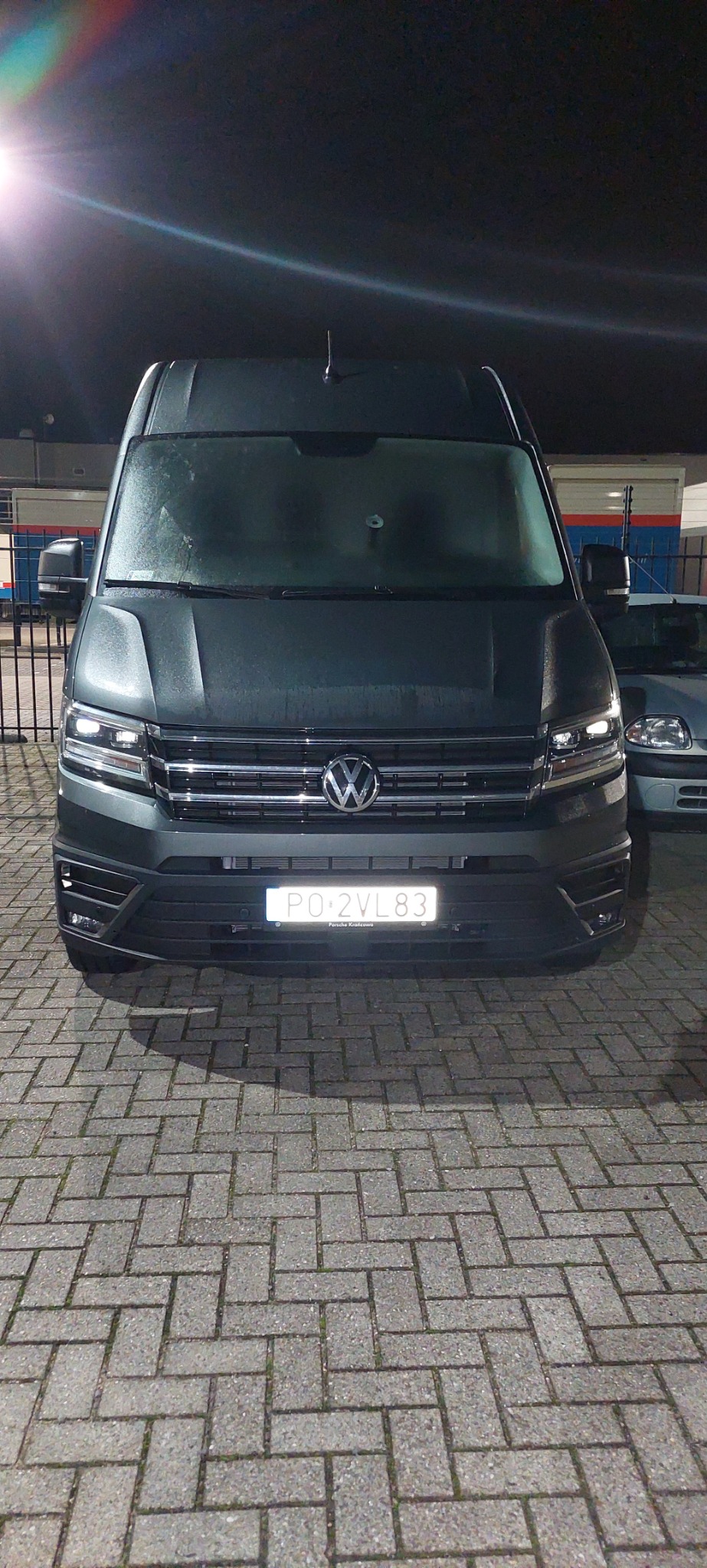 Czarny Volkswagen Crafter z polską tablicą rejestracyjną PO 2VL83 zaparkowany na brukowanym placu, widok z przodu, oświetlony światłem lampy ulicznej.