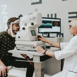 Pacjent podczas badania wzroku za pomocą urządzenia Topcon 3D OCT-1 Maestro, obsługiwanego przez okulistkę w białym fartuchu, z tablicą Snellena w tle.