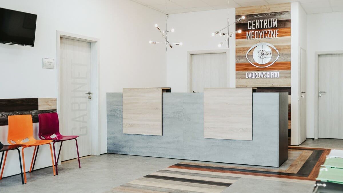 Recepcja w centrum medycznym z logo oka na drewnianej ścianie, nowoczesne lampy i kolorowe krzesła dla pacjentów.