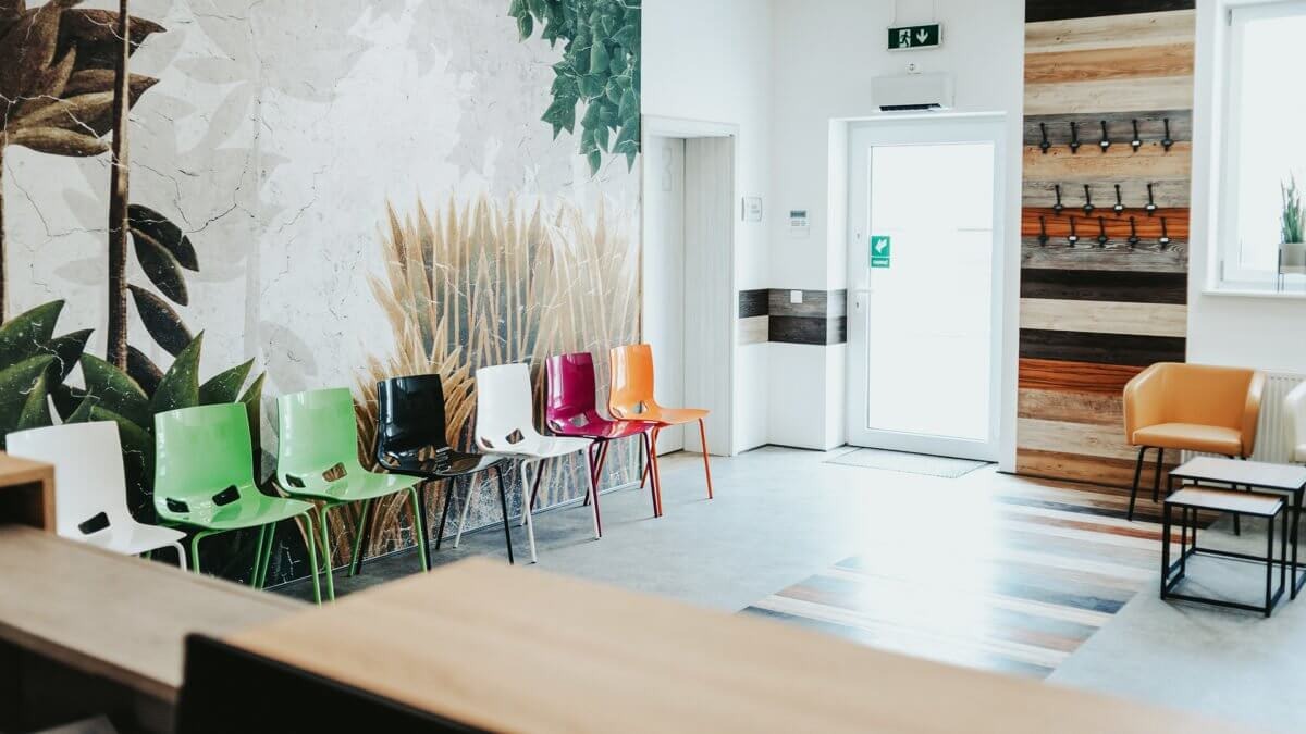 Poczekalnia w gabinecie lekarskim z rzędem kolorowych krzeseł na tle ściany z motywem roślinnym i drewnianym panelem z wieszakami.