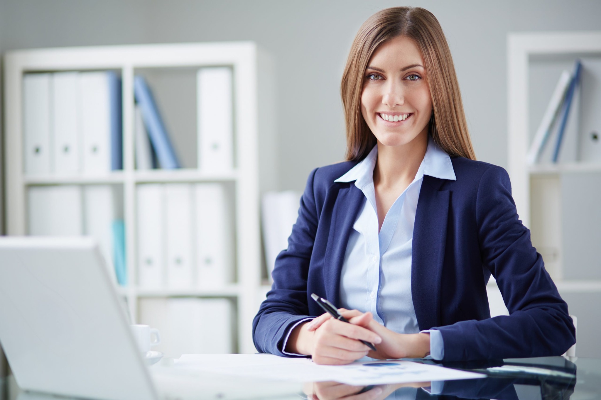 Uśmiechnięta kobieta w biurze, ubrana w granatową marynarkę i jasną koszulę, trzyma długopis nad dokumentami, na biurku laptop i segregatory w tle.