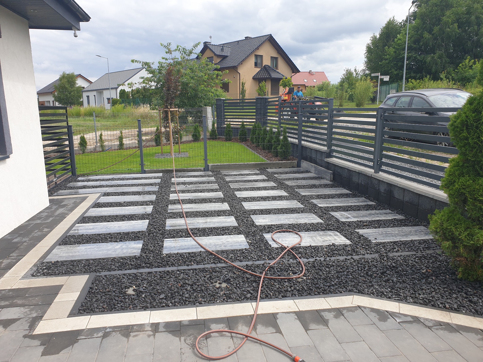 Nowoczesny podjazd z prostokątnych płyt ułożonych w szachownicę, wypełniony czarnym grysem, z widocznym wężem ogrodowym i fragmentem ogrodzenia.