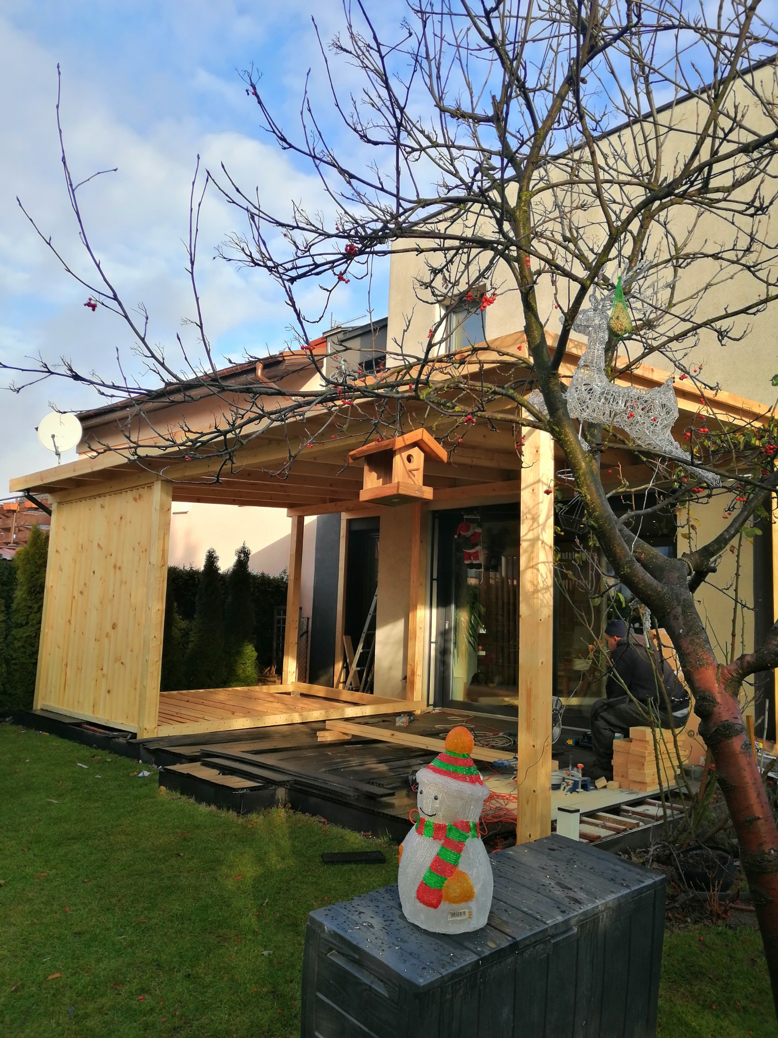 Drewniana konstrukcja zadaszenia tarasu w trakcie budowy, widoczny częściowo ułożony drewniany podest, obok drzewo z dekoracją świetlną renifera i plastikowy bałwanek, w tle dom jednorodzinny.