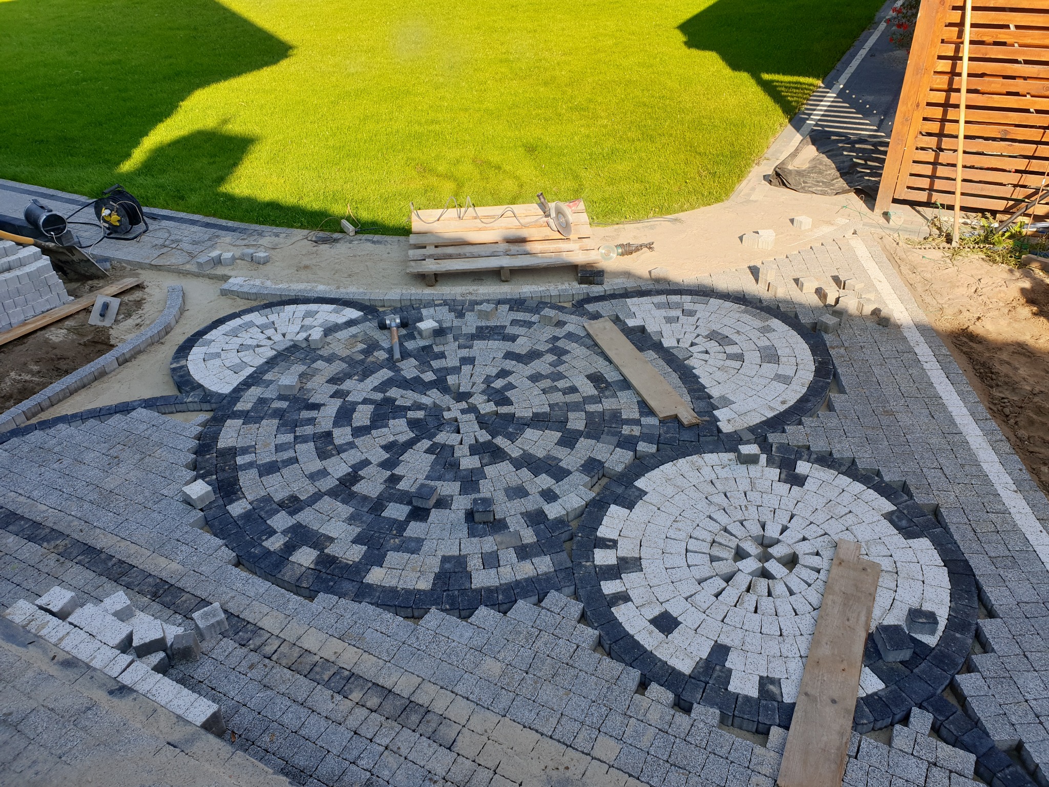 Fragment podjazdu z szarej kostki brukowej układanej we wzór koncentrycznych okręgów z użyciem kostki w dwóch odcieniach szarości, widoczne narzędzia i materiały budowlane w tle.
