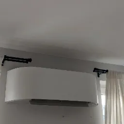 Biały klimatyzator ścienny zamontowany w pokoju z jasnymi zasłonami i czarnymi karniszami, widok z dołu.