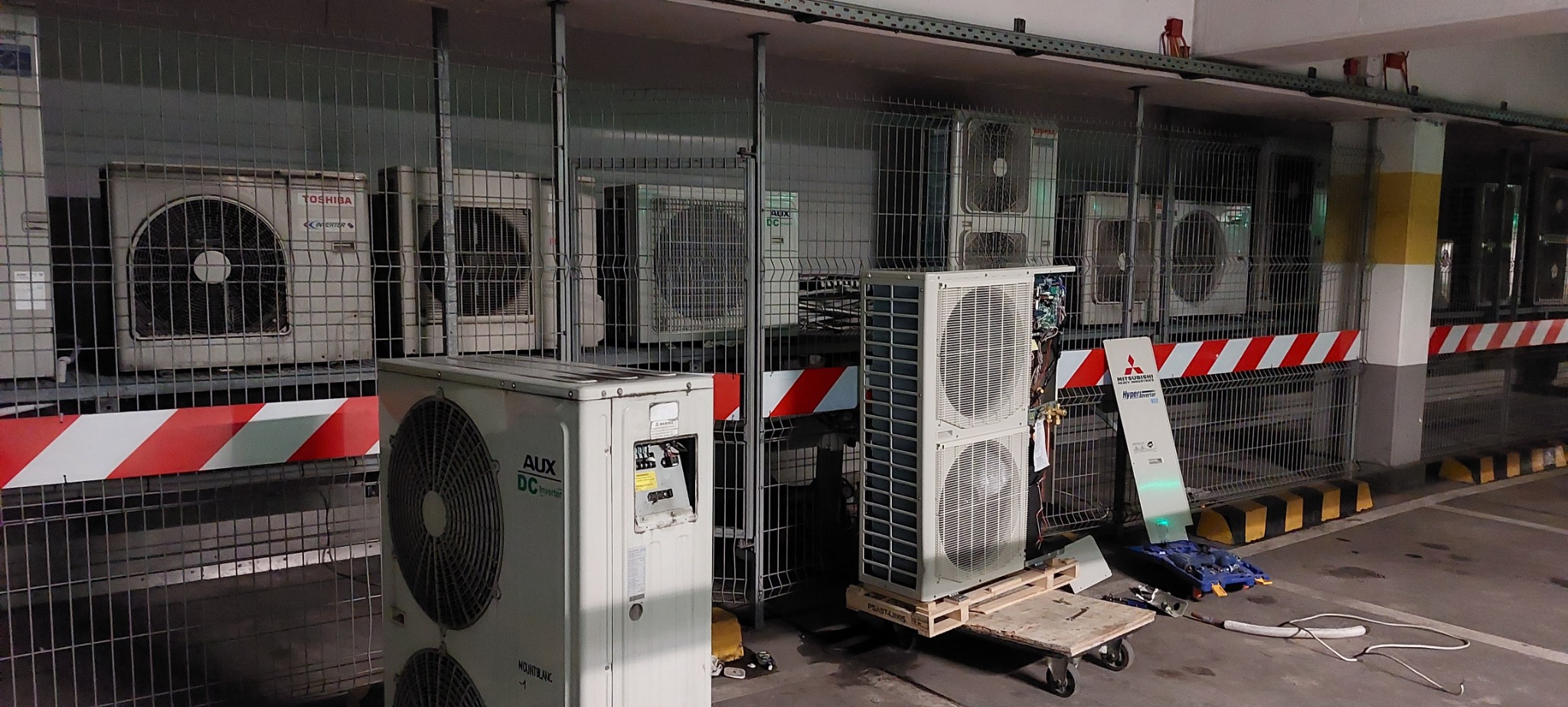 Kolekcja jednostek zewnętrznych klimatyzacji różnych marek (Toshiba, AUX, Mitsubishi) w garażu, część na paletach, jedna z otwartą obudową.