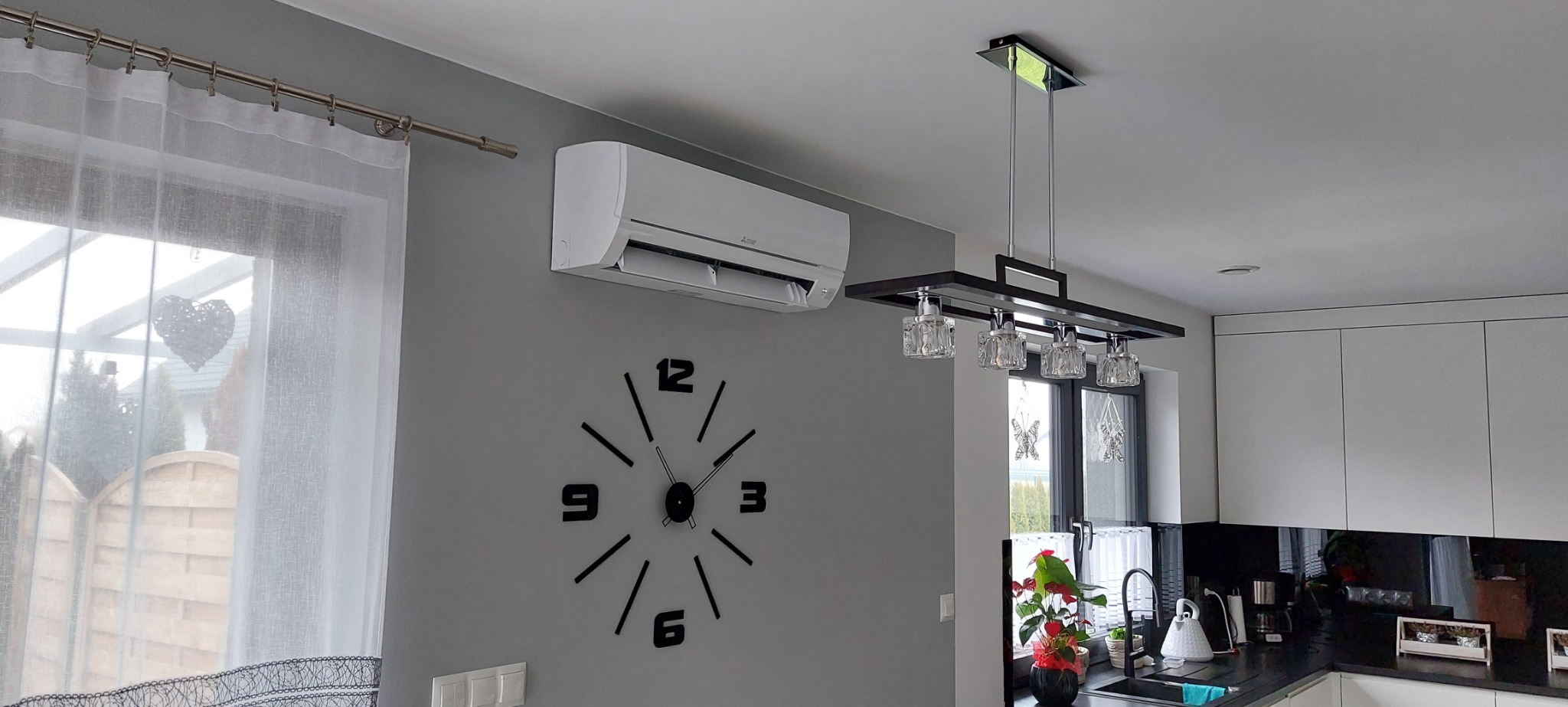Biały klimatyzator Mitsubishi zamontowany na szarej ścianie w nowoczesnej kuchni, obok dekoracyjny zegar i designerska lampa wisząca.