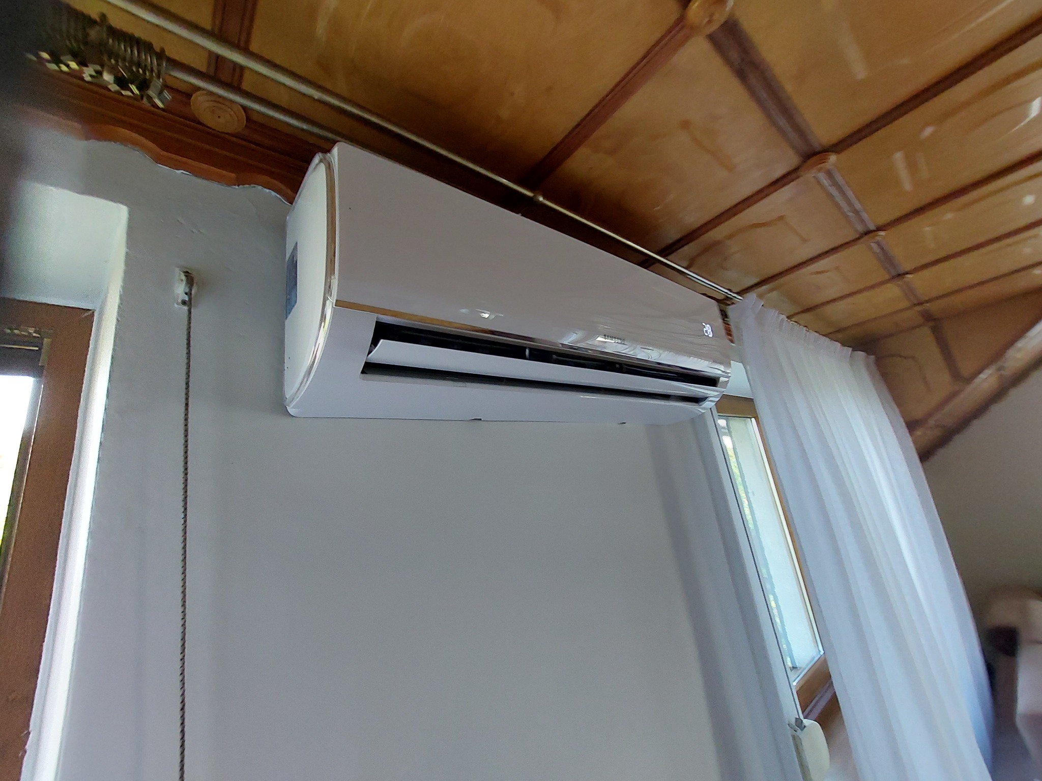 Biały klimatyzator ścienny zamontowany w pomieszczeniu z drewnianym sufitem i oknem zasłoniętym białą firanką, widok z dołu.