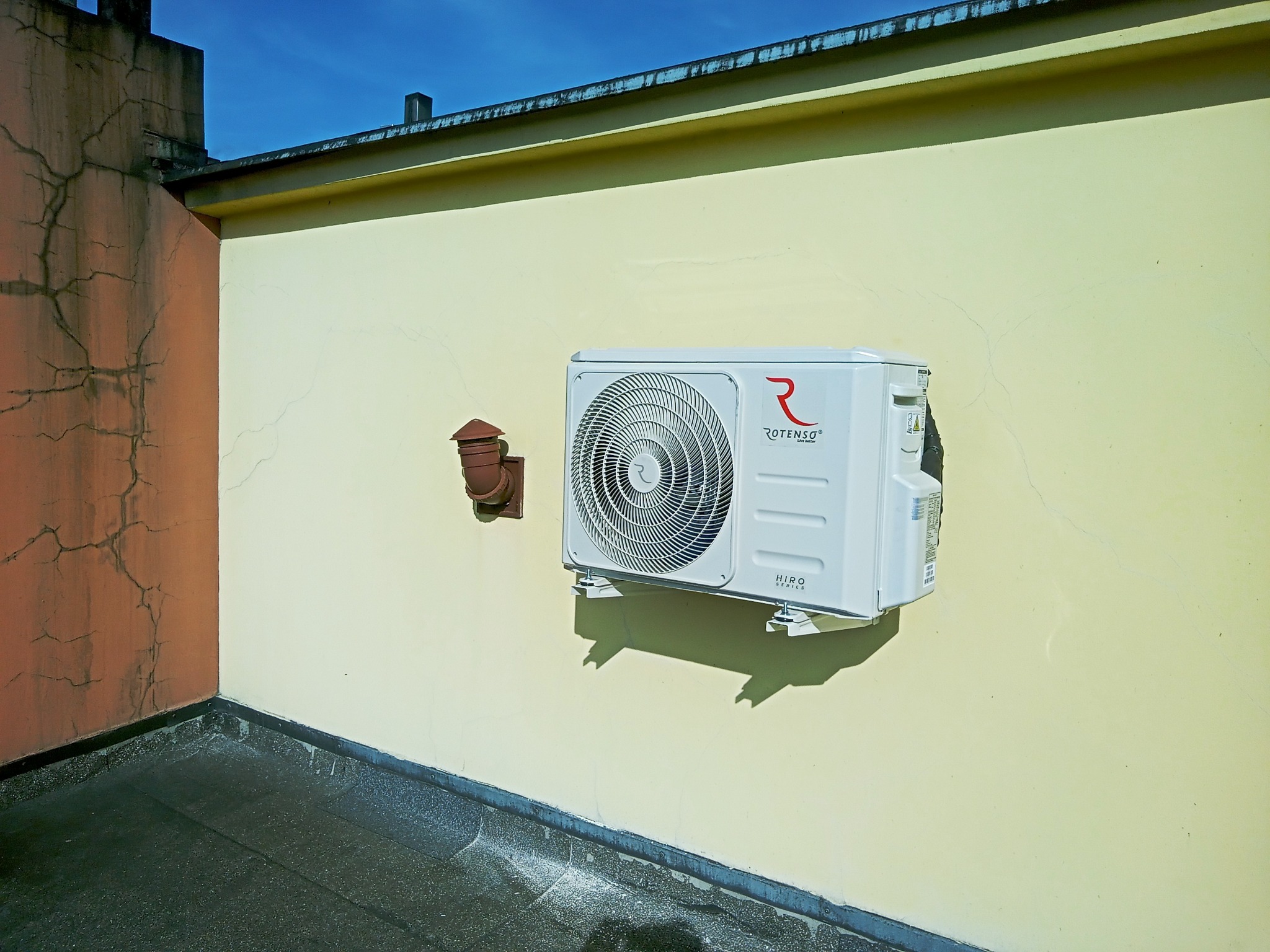 Zewnętrzna jednostka klimatyzacji Rotenso Hiro Series zamontowana na żółtej ścianie budynku, obok brązowej rury wentylacyjnej, widoczne pęknięcia na ścianie i fragment dachu.