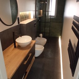 PROPAINT - Zajączkowski Jakub - Nowoczesna łazienka z drewnianym blatem pod umywalką nablatową, czarną armaturą, toaletą i bidetem w tle, podświetlona listwą LED wzdłuż ściany.