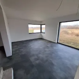 Nowoczesne wnętrze z szarą podłogą panelową i dużymi oknami z widokiem na krajobraz, widoczne kable wystające z sufitu.