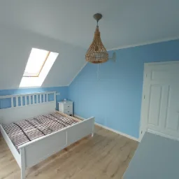 Pokój na poddaszu po remoncie: pomalowane na błękitno ściany, białe meble, drewniana podłoga, okno dachowe, pleciona lampa wisząca.