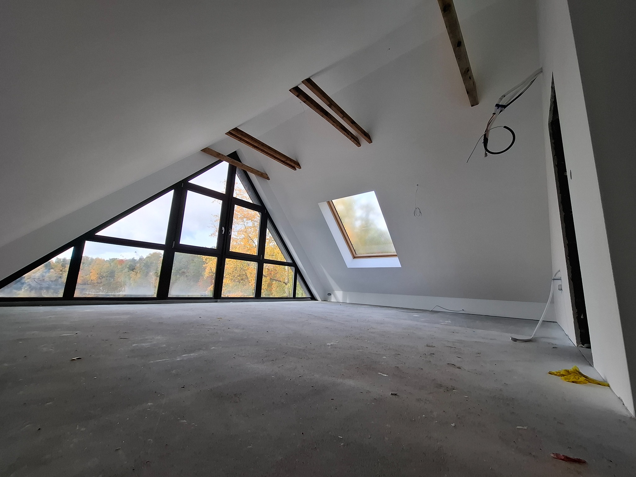 Poddasze w trakcie prac wykończeniowych, widoczne okno trójkątne z widokiem na jesienne drzewa, okno dachowe, surowe drewniane belki i przewody elektryczne.