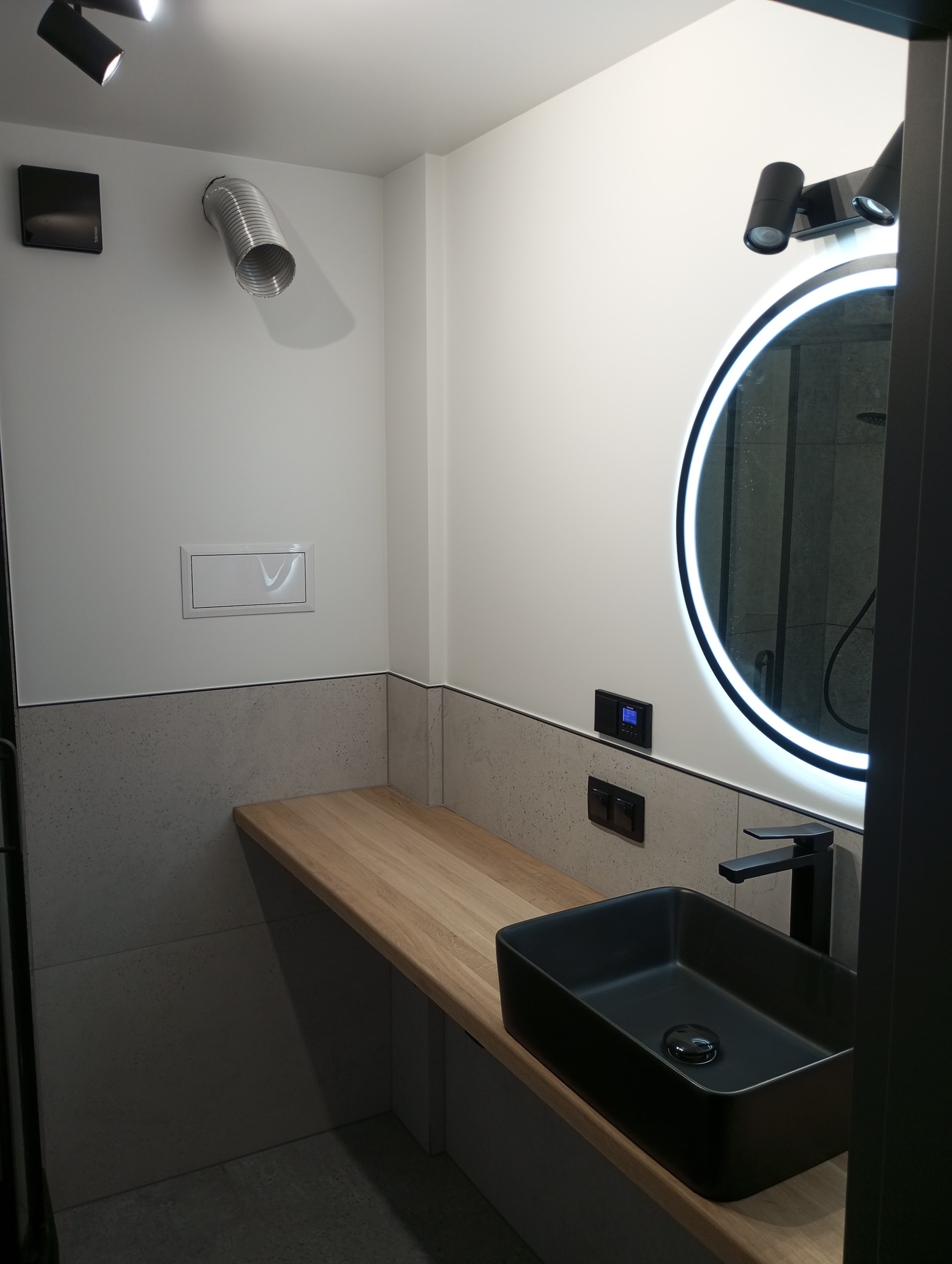 Nowoczesna łazienka z czarną, prostokątną umywalką nablatową, drewnianym blatem, okrągłym lustrem z podświetleniem LED i szarą płytką imitującą beton na ścianie, oświetlona punktowymi reflektorami.